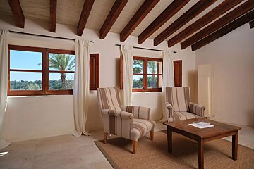 A rustic living room with wooden beamed ceiling, two striped armchairs, a coffee table, and windows revealing palm trees and greenery outside.