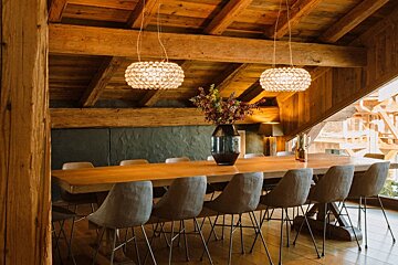 A cozy dining room featuring a long wooden table, numerous grey chairs, a beamed ceiling, and two ornate crystal chandeliers illuminating a floral centerpiece.