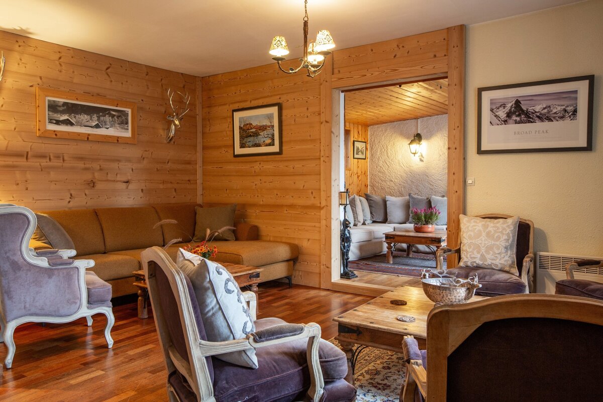 A rustic, wood-paneled living room features a brown sectional, purple armchairs, wooden tables, and framed art. An open doorway leads to another cozy sitting area.