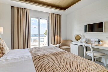 Bright hotel room with a bed, desk, TV, and a balcony offering a beautiful sea view.