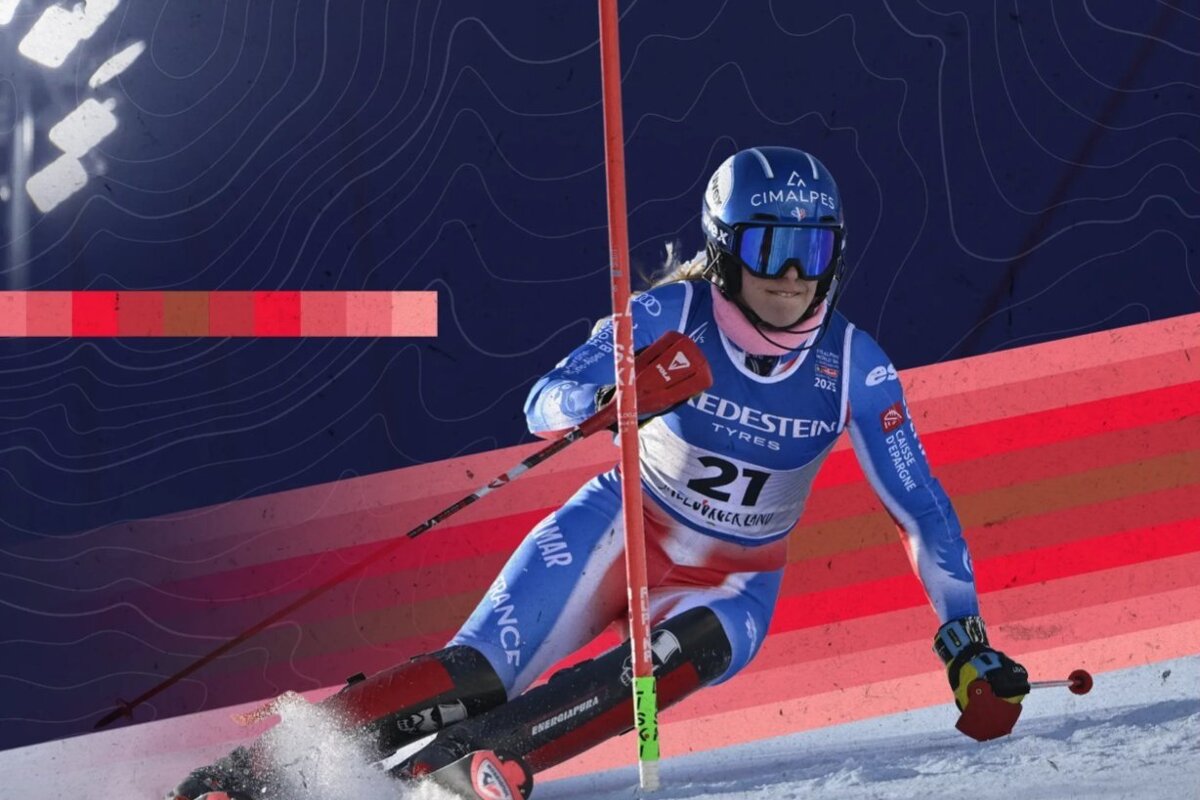 A female alpine skier in a blue suit and helmet navigates a course, kicking up snow. The background features blue contour lines and red stripes.
