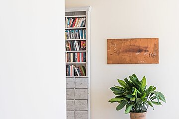 A minimalist interior featuring a book-filled shelf, a textured brown painting, and a vibrant potted plant against light walls.