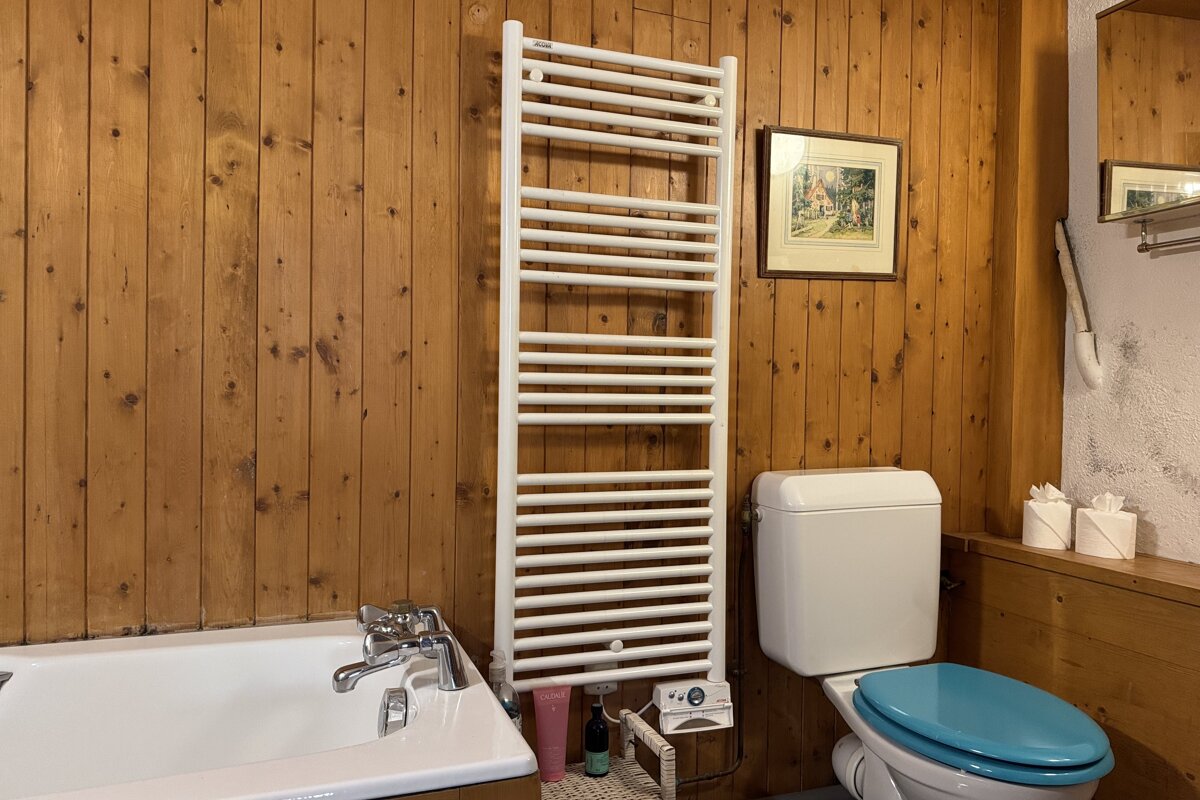 A wood-paneled bathroom with a white tub, tall towel warmer, and toilet with a blue seat. A framed picture is on the wall.