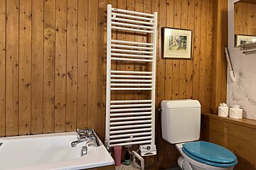 A wood-paneled bathroom with a white tub, tall towel warmer, and toilet with a blue seat. A framed picture is on the wall.