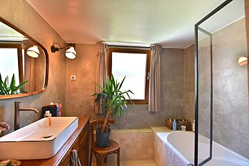 A modern bathroom with textured walls, a long white sink on a wooden vanity, a bathtub with a glass screen, a window, and a large plant.
