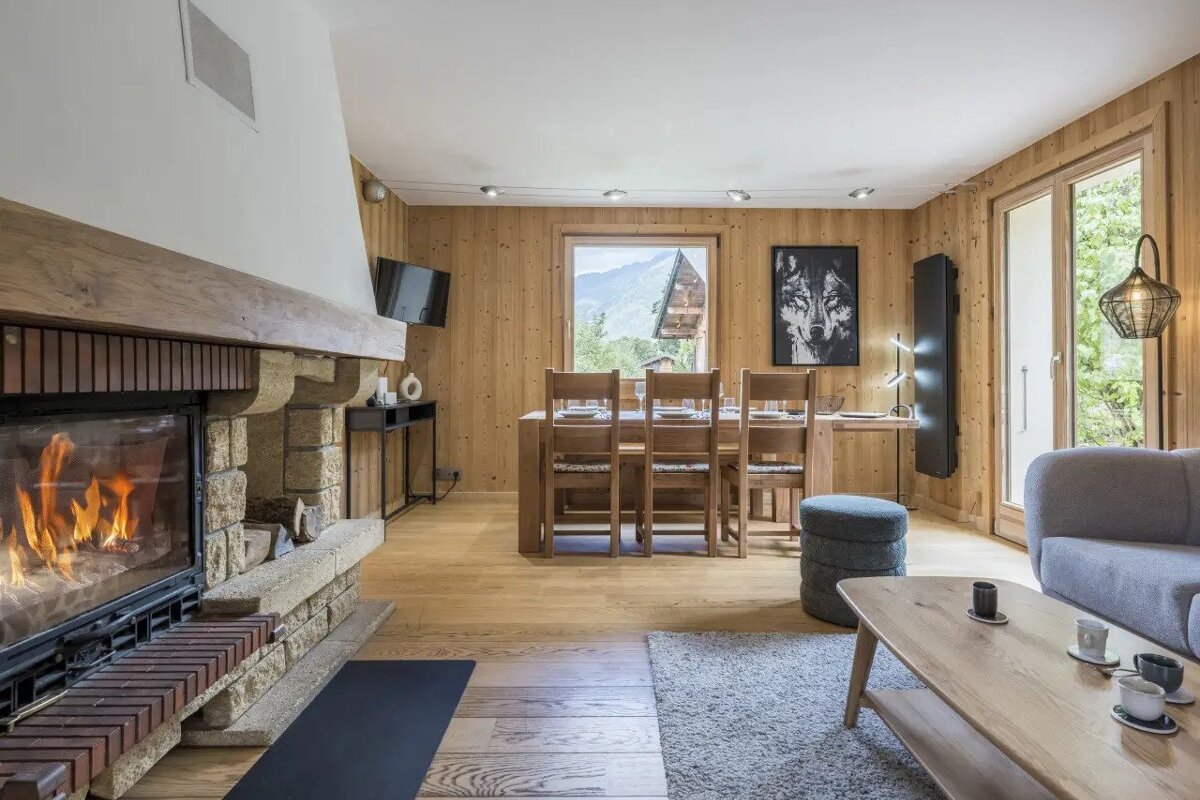 A cozy chalet living room with a roaring fireplace, wooden walls & floors, mountain views, a dining area, and a large wolf portrait.