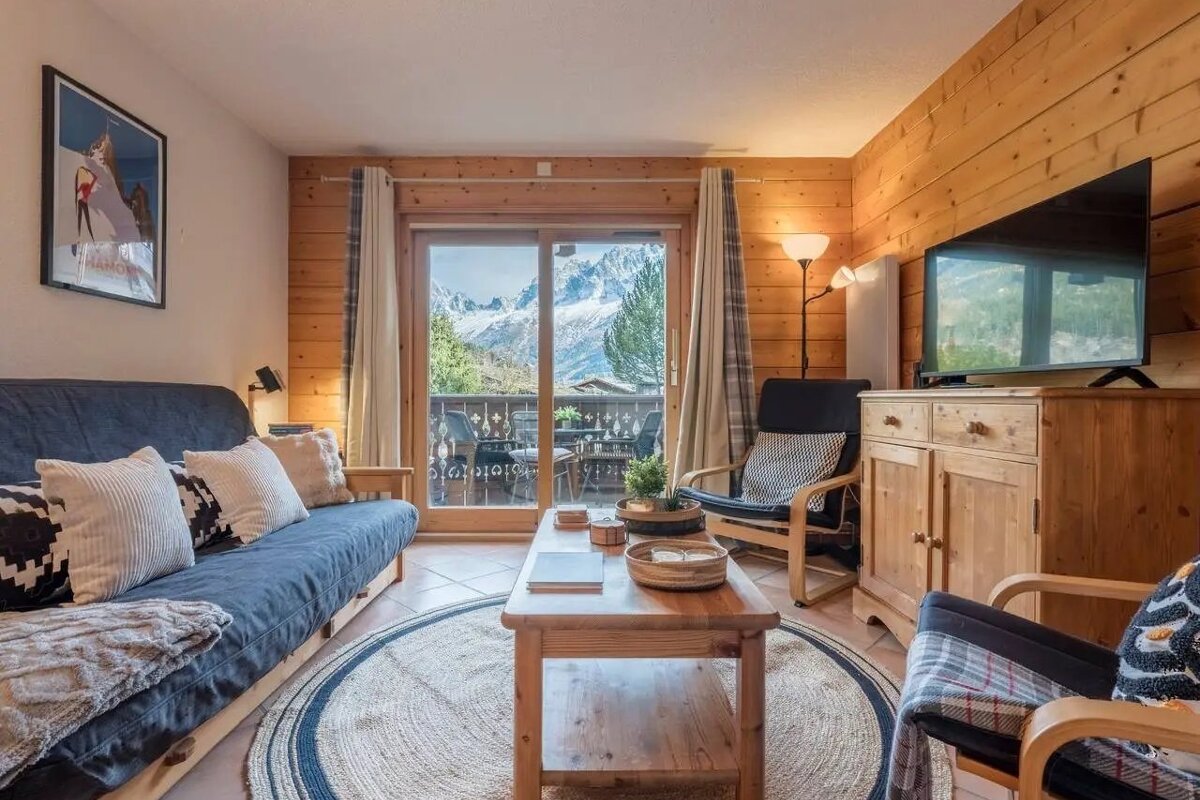 Cozy wood-paneled living room with a sofa, TV, and coffee table, opening to a balcony with a stunning view of snowy mountains.