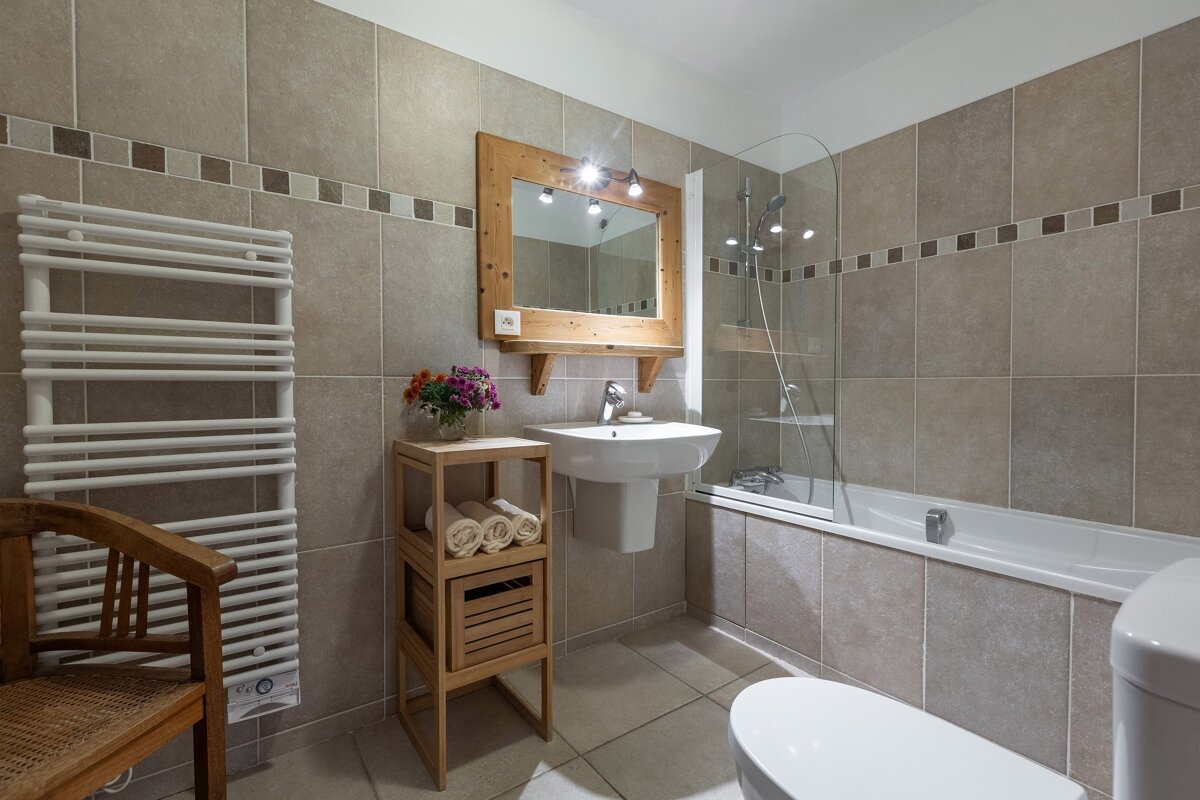 A warm-toned bathroom with beige tiles, featuring a bathtub with shower screen, a sink with a rustic wooden mirror, toilet, towel warmer, and a wooden storage unit with rolled towels and flowers.