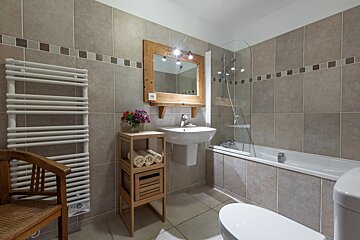 A warm-toned bathroom with beige tiles, featuring a bathtub with shower screen, a sink with a rustic wooden mirror, toilet, towel warmer, and a wooden storage unit with rolled towels and flowers.