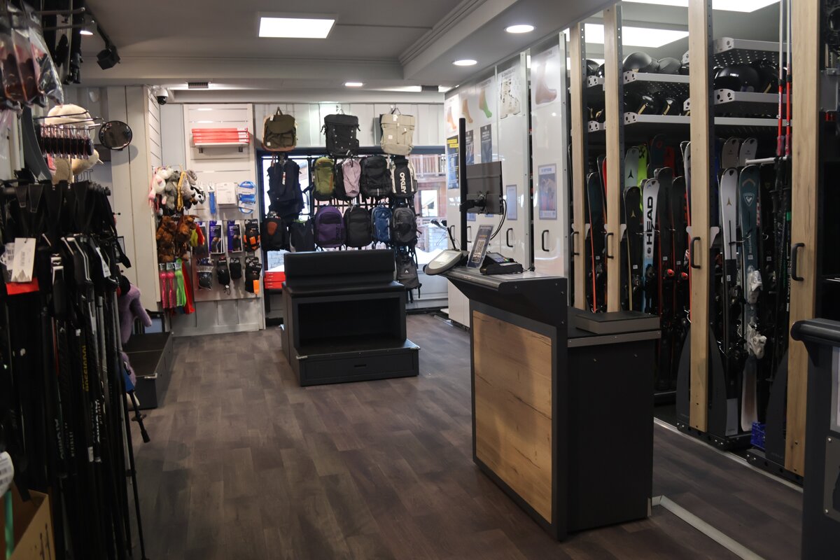 A well-lit ski shop interior featuring displays of skis, poles, helmets, backpacks, and other winter sports accessories, with a cash register.
