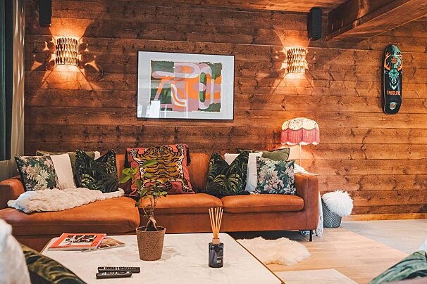 A cozy, rustic living room with rich wood-paneled walls, a warm orange sofa, and eclectic cushions. Features soft lighting, modern art, and unique decor.