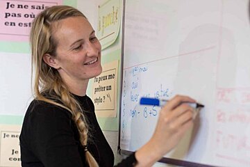A woman is writing on a whiteboard with a sign on the wall that says je ne sais pas ou en est