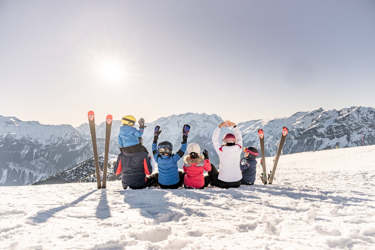 A family of five, backs to the camera, sits on a sunny snowy mountain, enjoying the vast mountain vista with skis propped up.