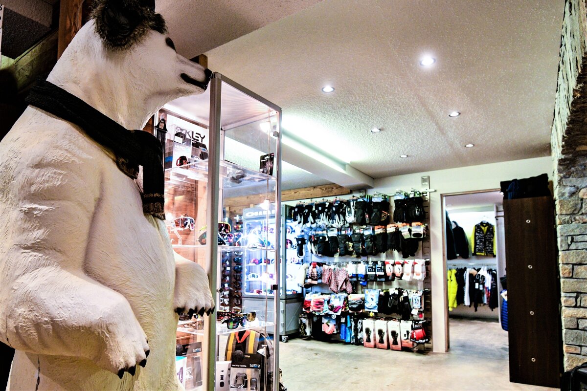 A large white polar bear statue wearing a scarf stands in a ski shop next to display cases. Racks of winter gloves, socks, and apparel fill the background.