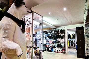 A large white polar bear statue wearing a scarf stands in a ski shop next to display cases. Racks of winter gloves, socks, and apparel fill the background.