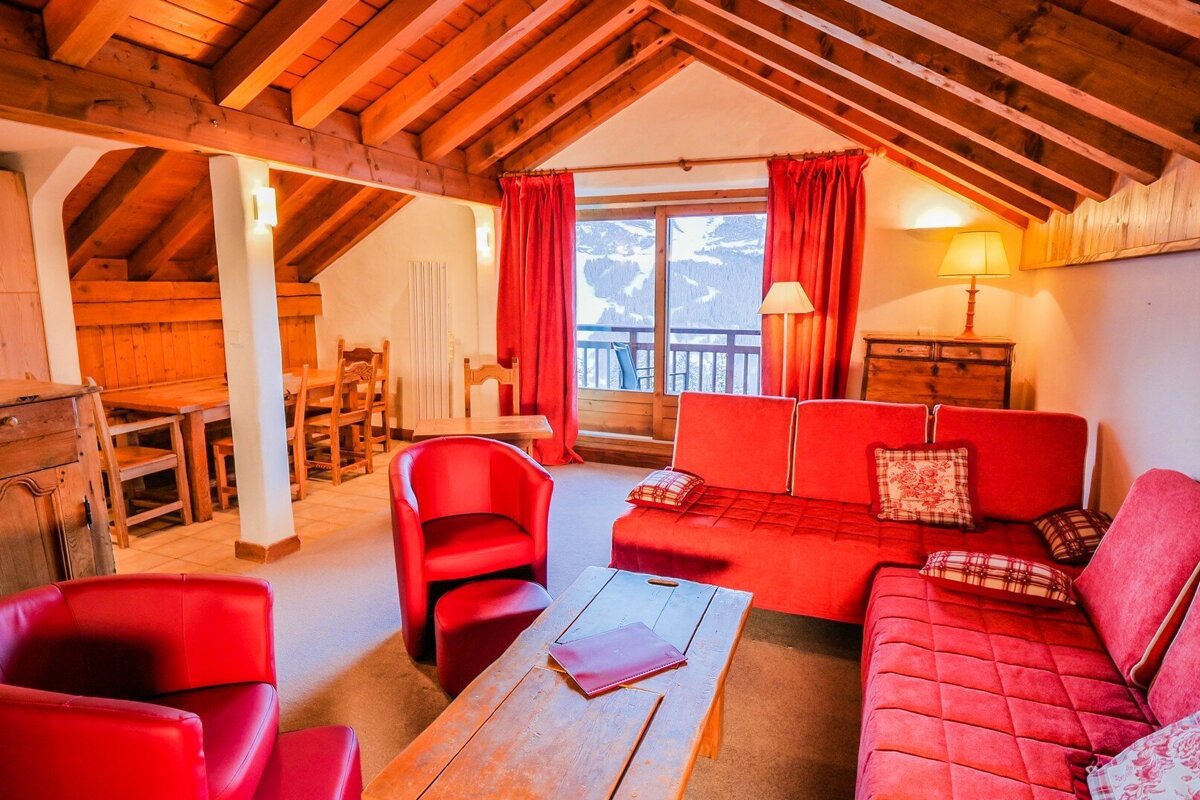 A cozy, rustic chalet living room with red sofas, armchairs, and curtains. It features a wooden beamed ceiling, dining area, and snowy mountain views.
