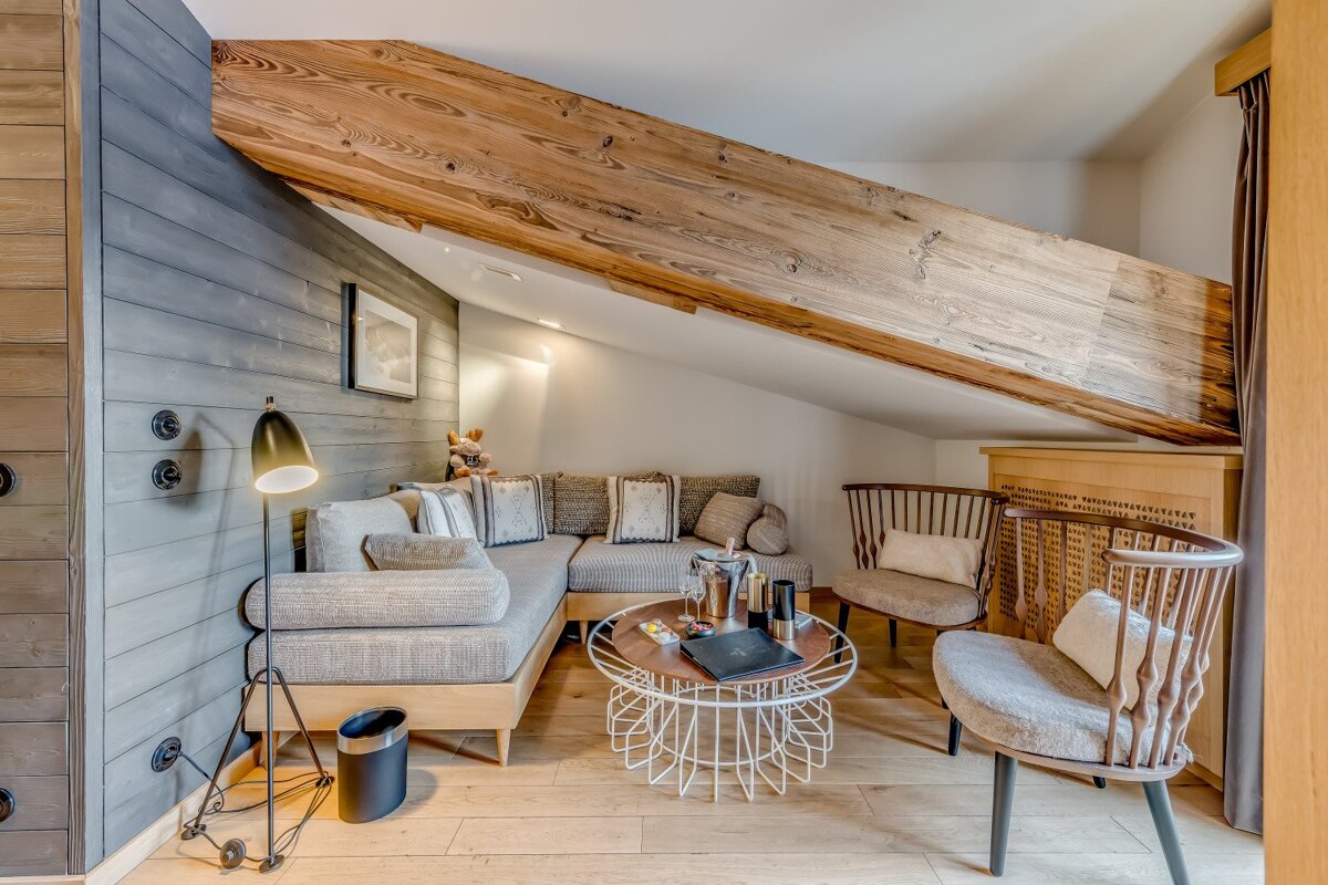 A cozy, modern chalet living room with an L-shaped sofa, two armchairs, and a coffee table under a prominent wooden beam and slanted ceiling.