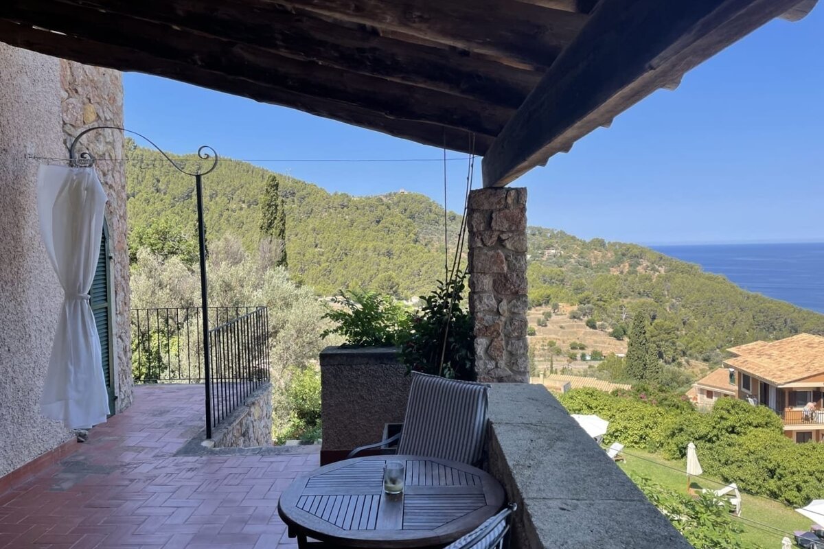 Scenic terraced balcony with table and chairs, offering panoramic views of forested mountains and the sparkling sea under a clear sky.