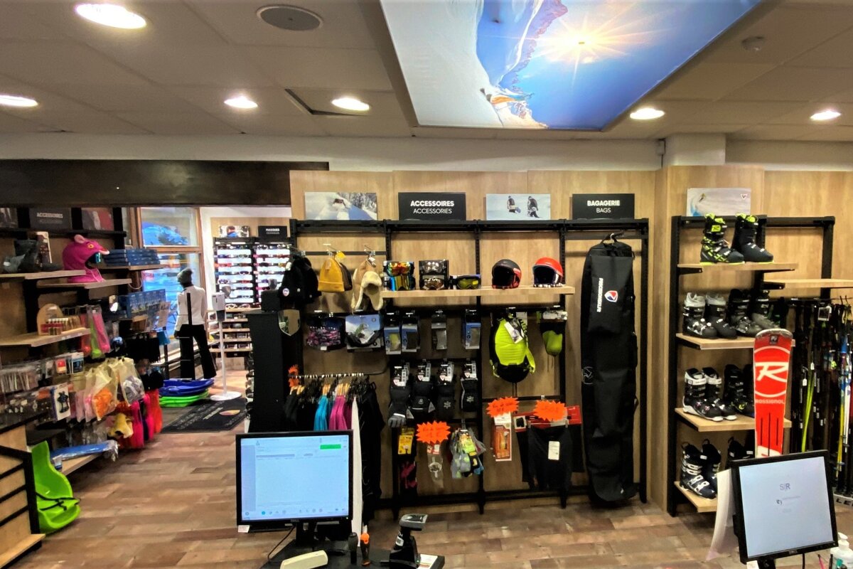 A brightly lit ski and winter sports shop featuring various gear like boots, skis, helmets, and accessories, with a large ceiling panel depicting a skier.