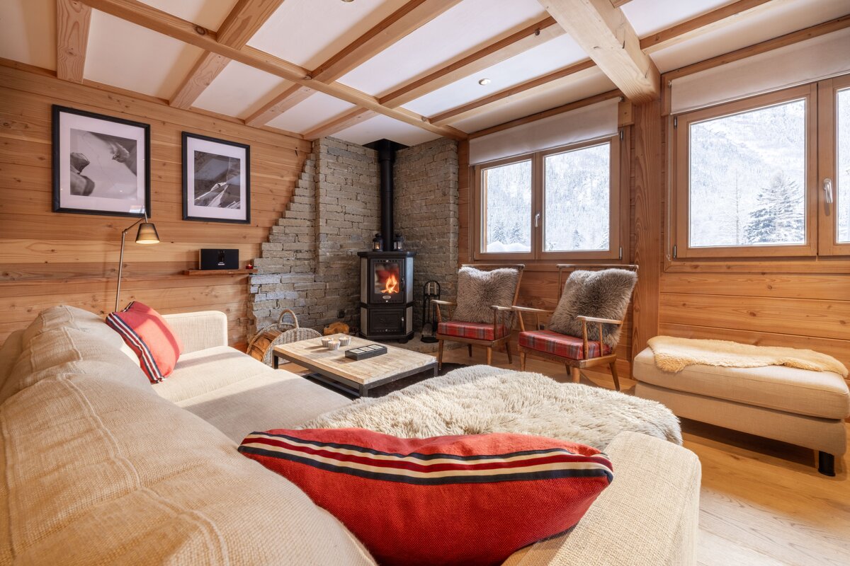A cozy, wood-paneled ski chalet living room with a crackling fireplace, plush seating, and snowy mountain views from the windows.