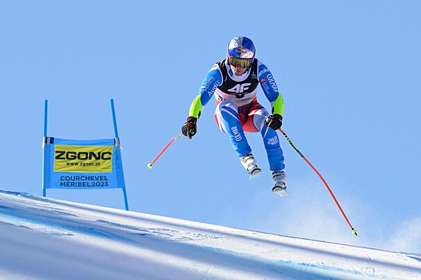 An alpine skier in a vibrant blue and red suit jumps high above a snowy slope during a race, under a clear blue sky.
