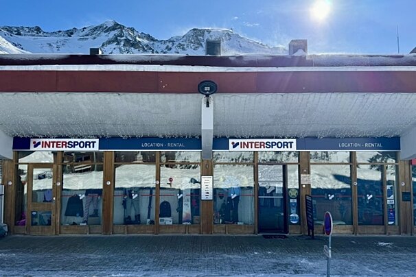 An Intersport ski rental shop in snowy mountains under a bright sun. Icicles adorn the roof, with ski gear visible inside.