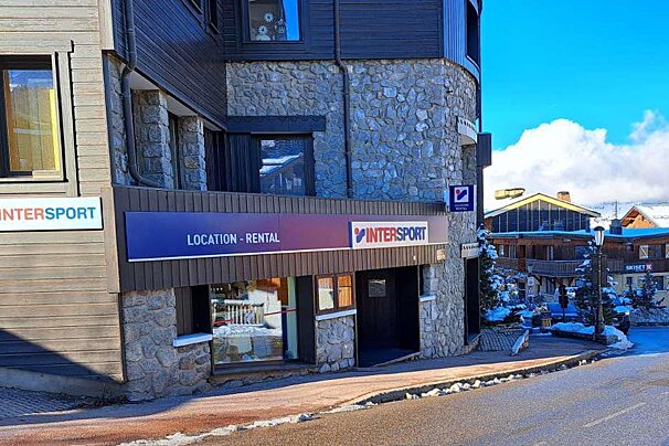 An Intersport rental shop with stone and wood architecture in a sunny, snowy mountain village, under a clear blue sky.