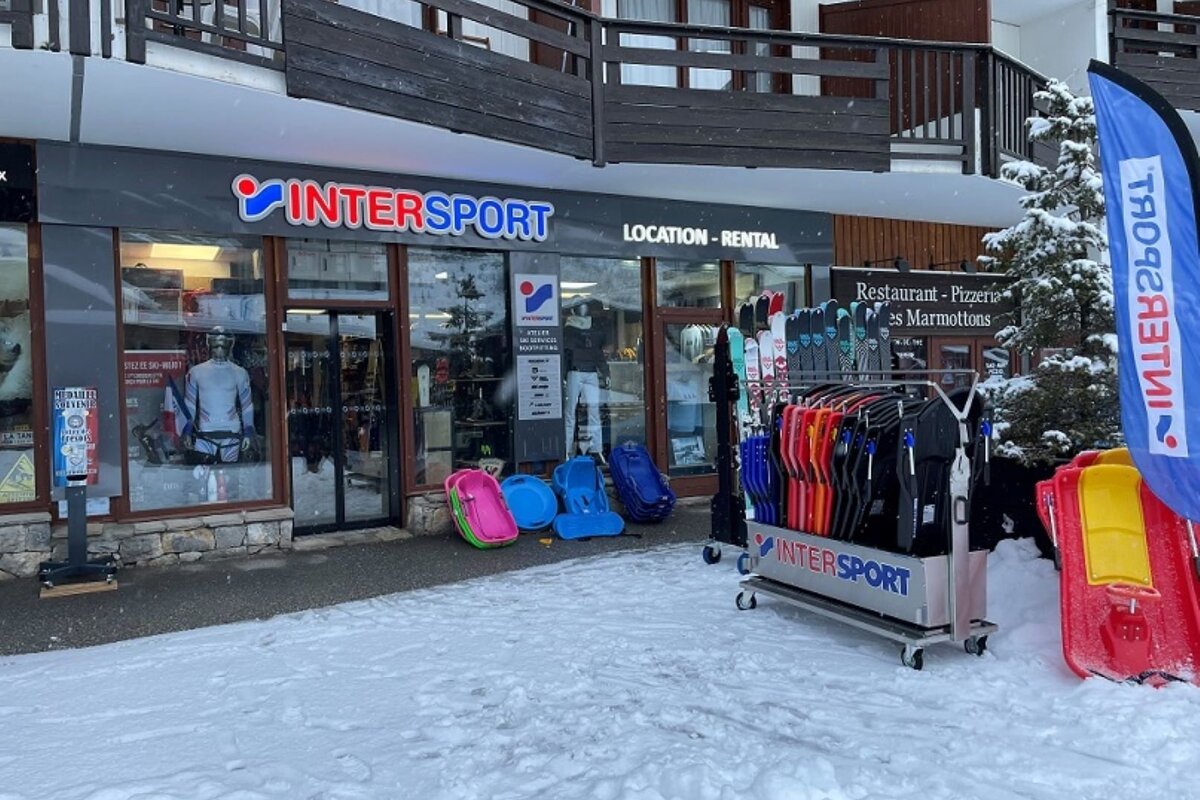 An Intersport ski rental shop in a snowy setting, displaying skis, snowboards, and sleds outside. The sign clearly reads 