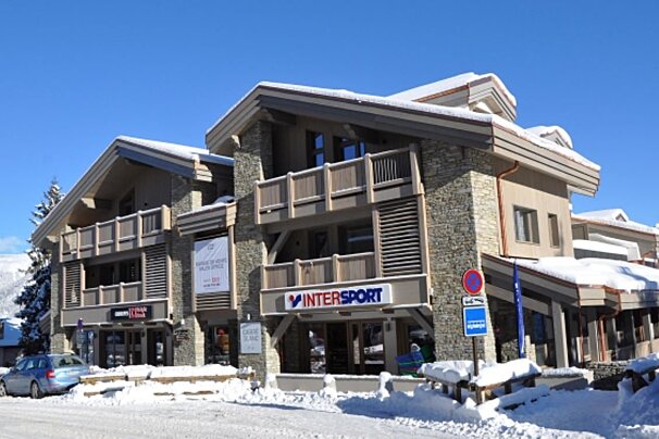 A multi-story stone and wood building, housing an Intersport store and other businesses, is covered in snow under a bright blue sky. A car is parked on the snowy street.