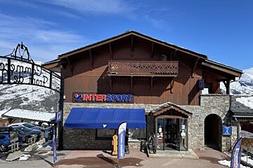 An Intersport ski shop in an alpine chalet building made of wood and stone, set against snowy mountains under a clear blue sky.