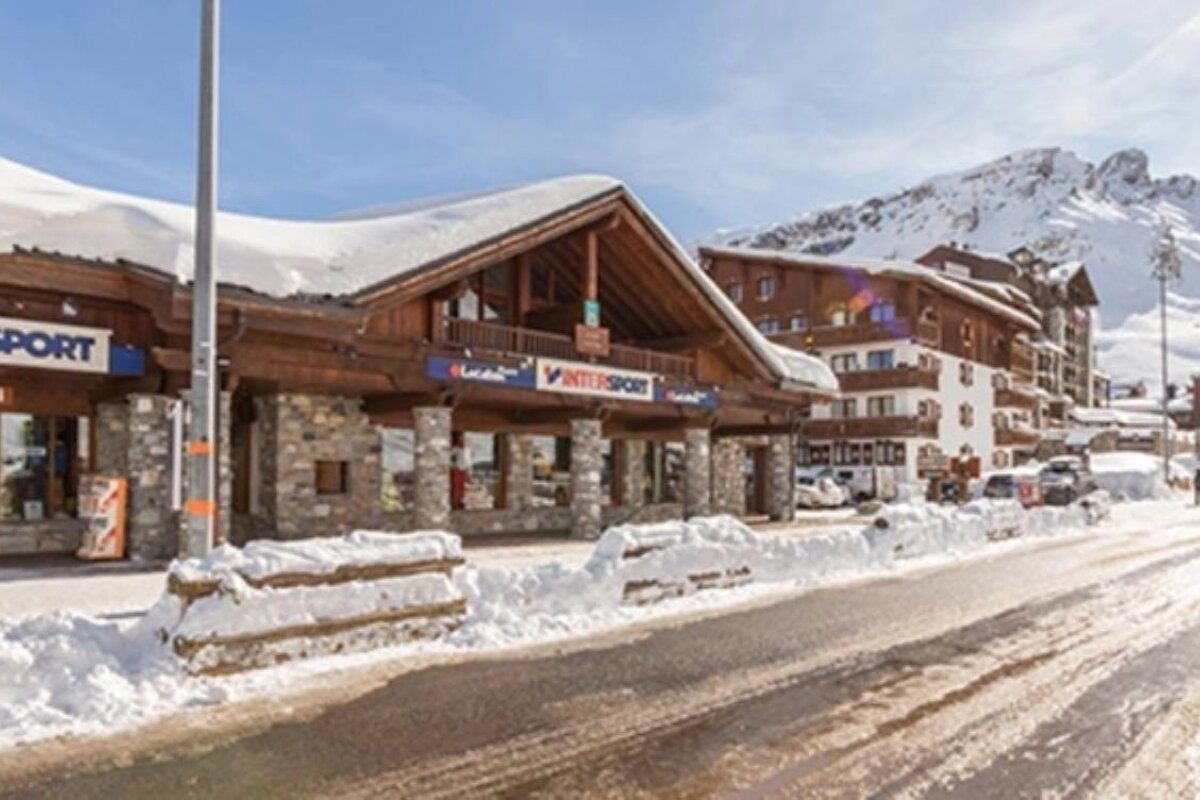A snowy mountain village street with wooden buildings, an Intersport store, a clear road, and distant snow-capped mountains under a blue sky.