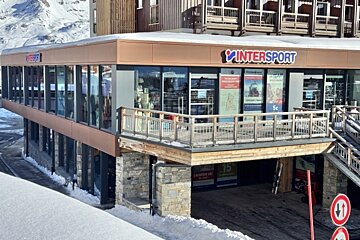 A modern, multi-level Intersport store is set in a snowy mountain resort, featuring large glass windows, a wooden balcony, and a stone base.