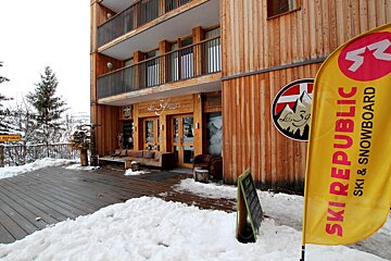A multi-story wooden building, likely a hotel or chalet, in a snowy winter setting. It features an outdoor terrace with seating and a 