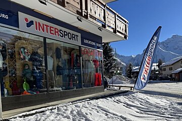 An Intersport ski shop front on a sunny, snowy day, displaying winter gear. Mountains loom in the background, and a blue flag stands outside.