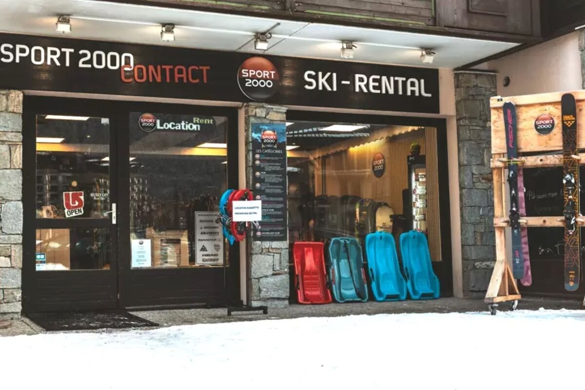 A Sport 2000 Ski-Rental shop entrance on a snowy day, displaying skis, snowboards, and red/blue sleds outdoors.