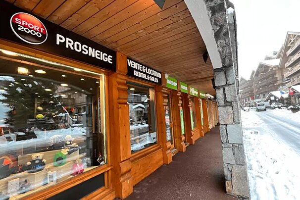 A snowy street in a mountain resort village, featuring a Sport 2000 Prosneige ski rental and sales shop with traditional wooden architecture.