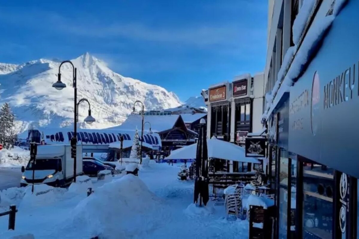 A snow-covered ski resort village street with buildings, streetlights, and towering mountains under a bright blue sky on a clear winter day.