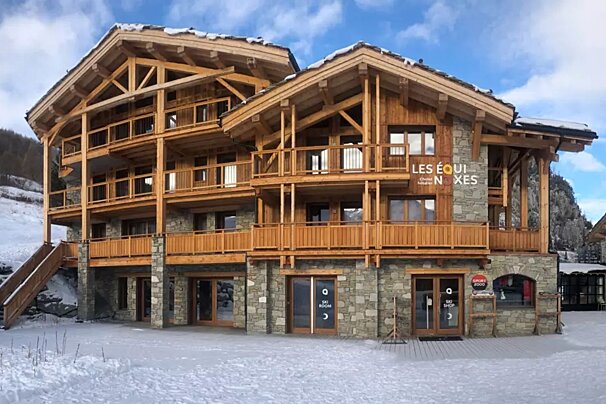 A large, multi-story ski chalet with stone and wood construction, balconies, a ski shop, and ski room, set in a snowy mountain landscape.
