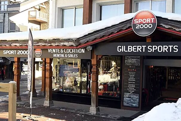 A snow-covered Sport 2000 / Gilbert Sports shop, advertising ski rental & sales. Winter sports gear is displayed inside the windows.