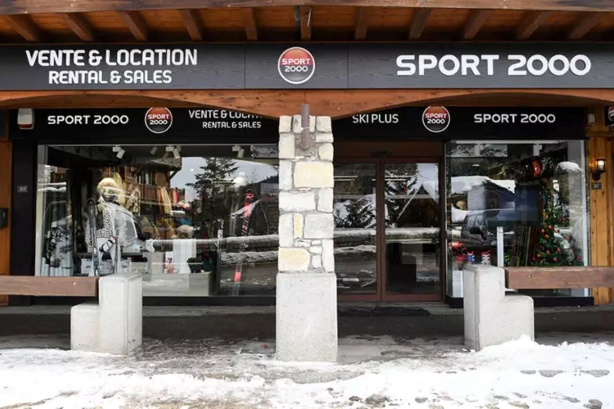 A Sport 2000 ski and rental shop storefront with snowy ground. Large windows display merchandise, reflecting a winter landscape and distant trees.