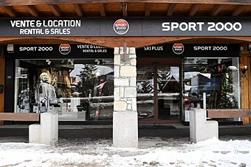 A Sport 2000 ski and rental shop storefront with snowy ground. Large windows display merchandise, reflecting a winter landscape and distant trees.