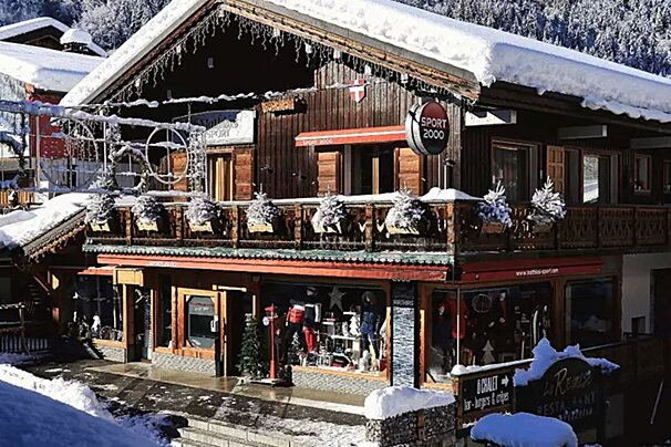 A snow-covered rustic wooden chalet houses a Sport 2000 ski shop. Icicles hang from the roof, adding to the charming winter mountain village scene.