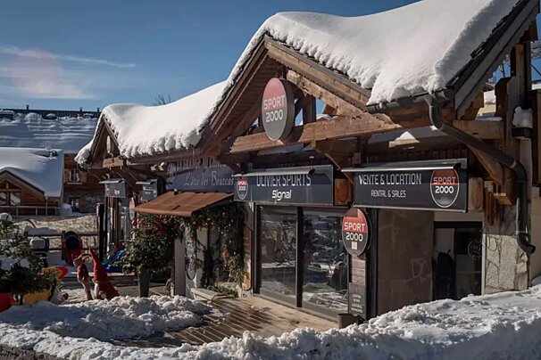 A snow-covered Sport 2000 shop (Olivier Sports, Rental & Sales) in a sunny, wintry mountain village, with festive decorations visible.