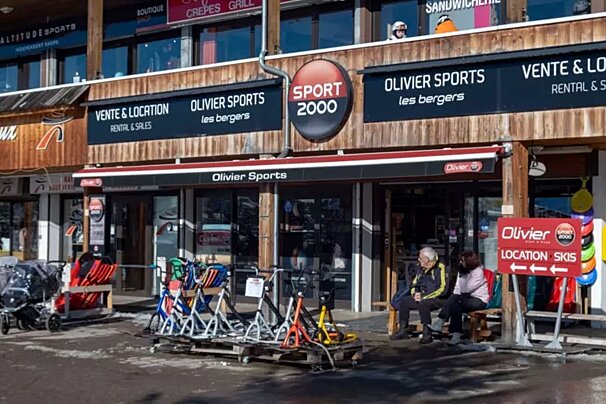 An Olivier Sports (Sport 2000) ski rental and sales shop. Equipment is displayed outside, and two people are seated on a bench.