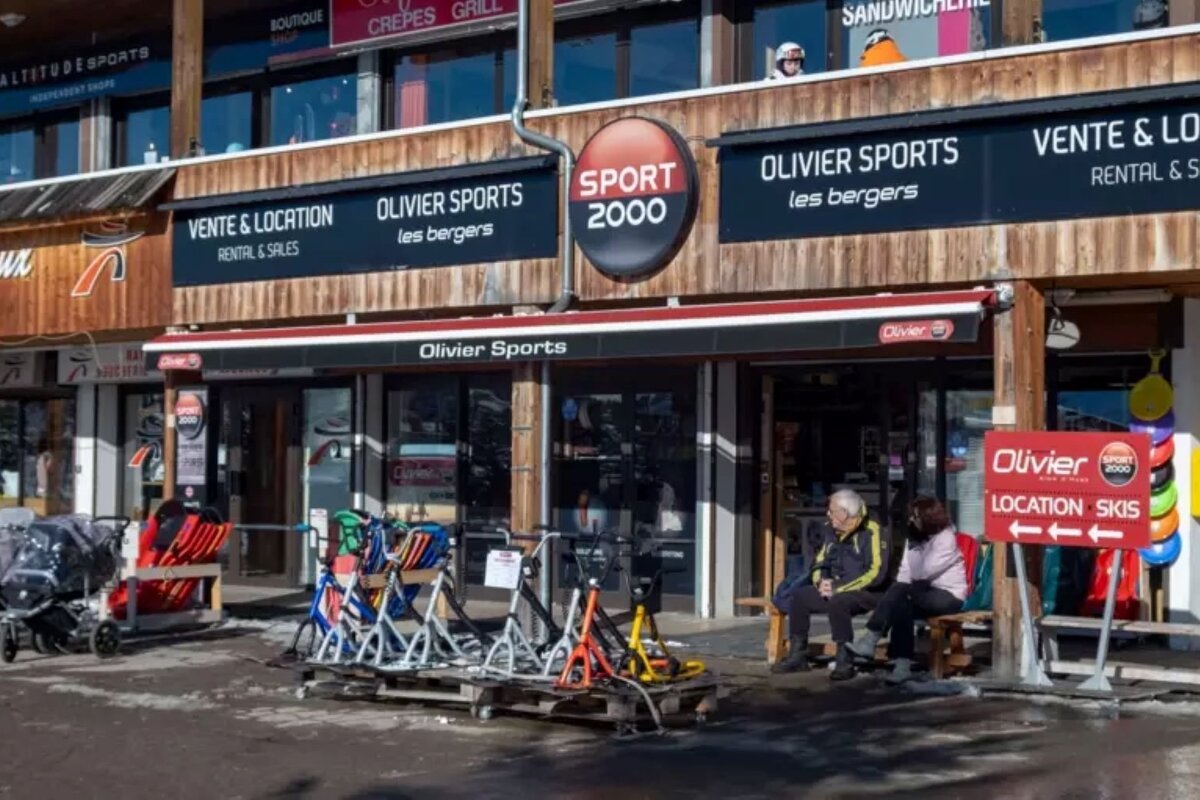 An Olivier Sports (Sport 2000) ski rental and sales shop. Equipment is displayed outside, and two people are seated on a bench.