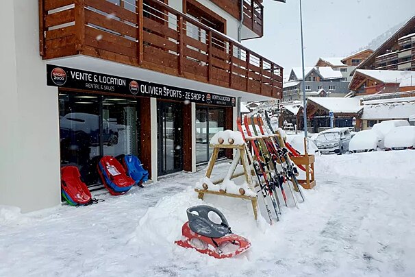 A snowy winter scene outside a ski rental shop (Olivier Sports) with skis, sleds, and snow-covered cars, suggesting a mountain resort.