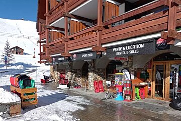 A snow-covered ski resort building with Sport 2000 rental shops on the ground floor, against a backdrop of snowy mountains and ski lifts.