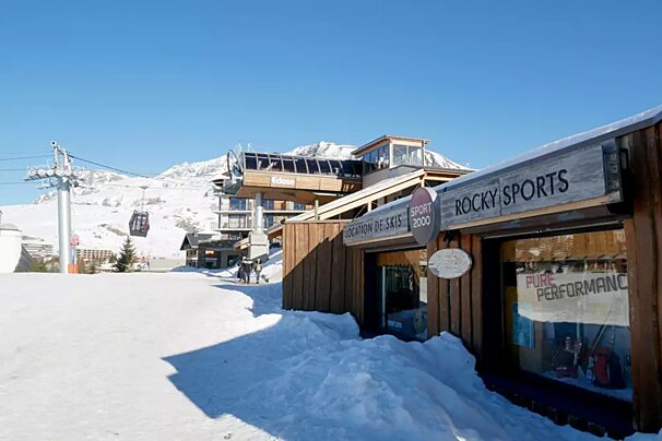 A sunny, snowy mountain resort scene with a ski lift, buildings, and a 