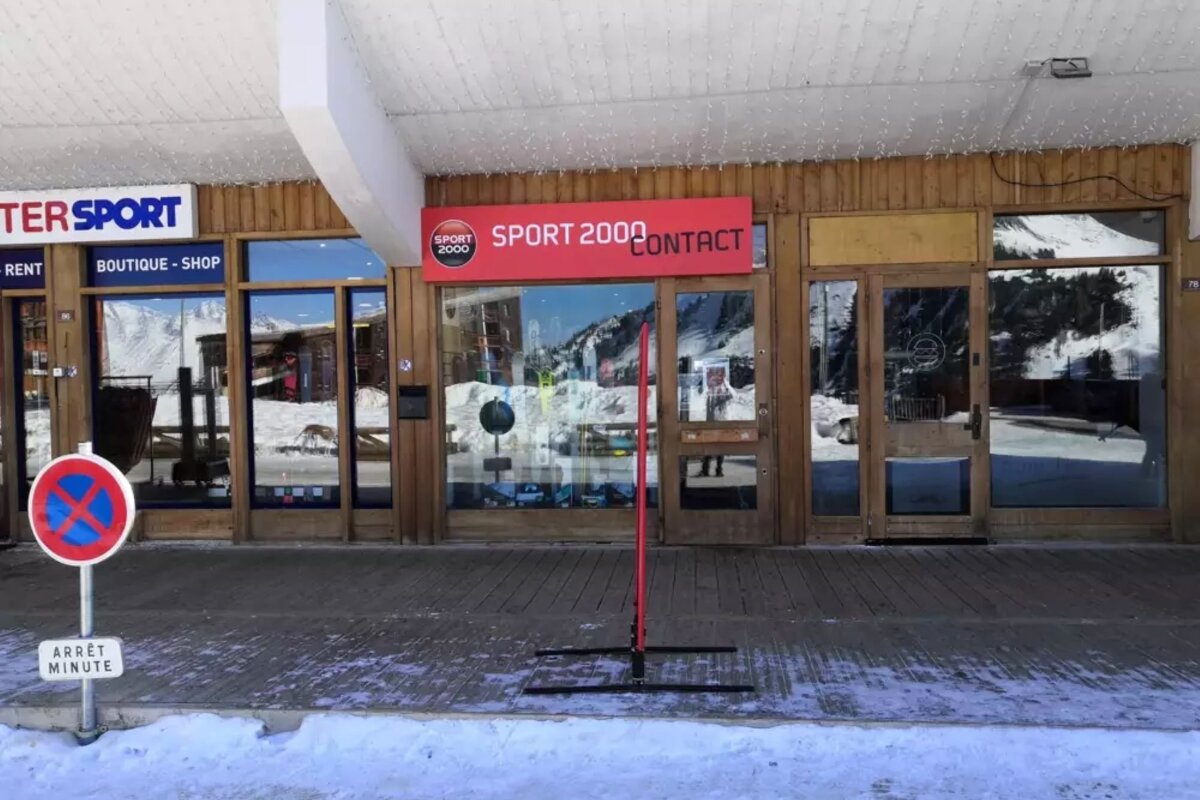 A snowy street in front of ski shops (Sport 2000, Intersport) with wooden facades and mountain reflections. A 