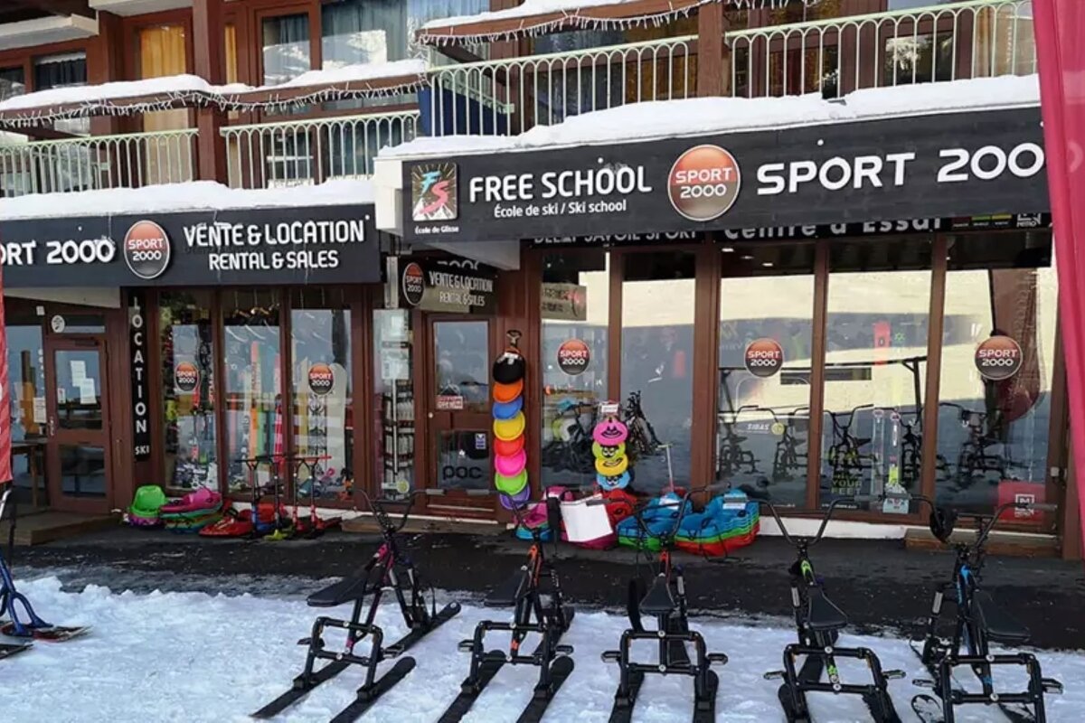 A snowy Sport 2000 ski rental and school with various winter sports equipment, including snow bikes, displayed outside in a mountain resort.
